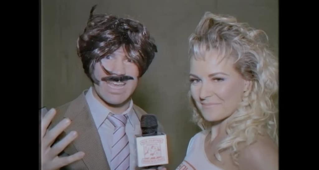 Renee Young as backstage interviewer Susan holding a microphone next to a heavily disguised Tyler Breeze as the evil banker Mr Mackelroy.