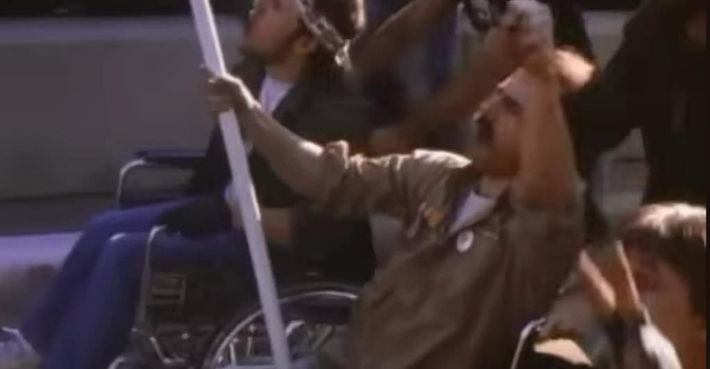 Tom Cruise portraying Ron Kovic in a wheelchair during a protest in orn on the Fourth of July (1989). Image credit: Born on the Fourth of July (1989) - film still via Rotten Tomatoes Classic Trailers/YouTube.