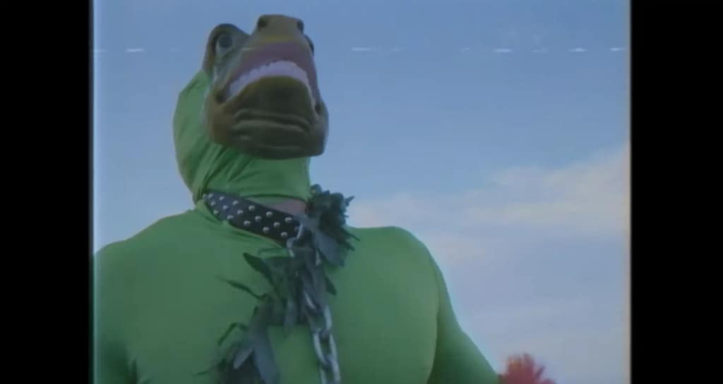 A low-angle shot of the Sea Creature character wearing a cheap green bodysuit and an oversized lizard mask against a blue sky.