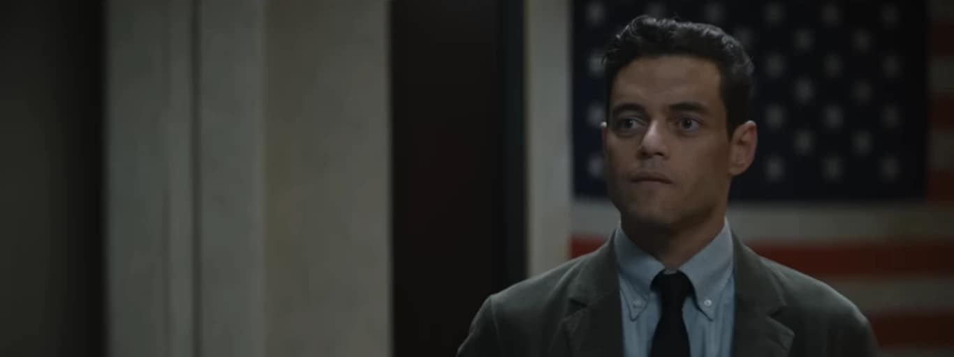 Charlie Heller (Rami Malek) standing in front of an American flag in The Amateur (2025).