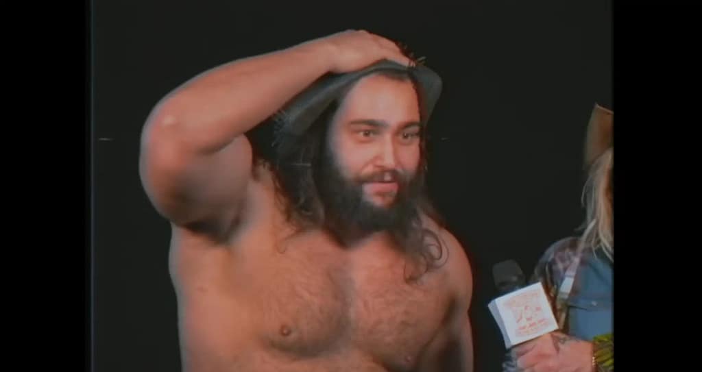 A close-up of a shirtless Rusev as the southern farmer Big Bartholomew clutching his hat in horror during a melodramatic promo segment.