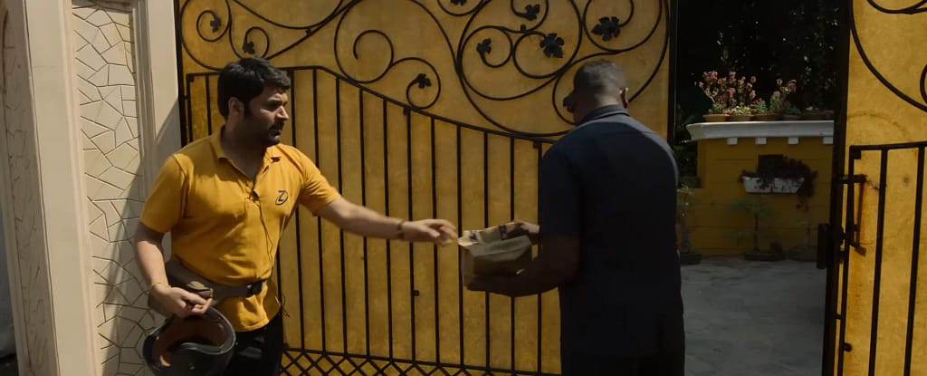 Manas (Kapil Sharma) handing over a food delivery package at a gated bungalow entrance in a scene from Zwigato.