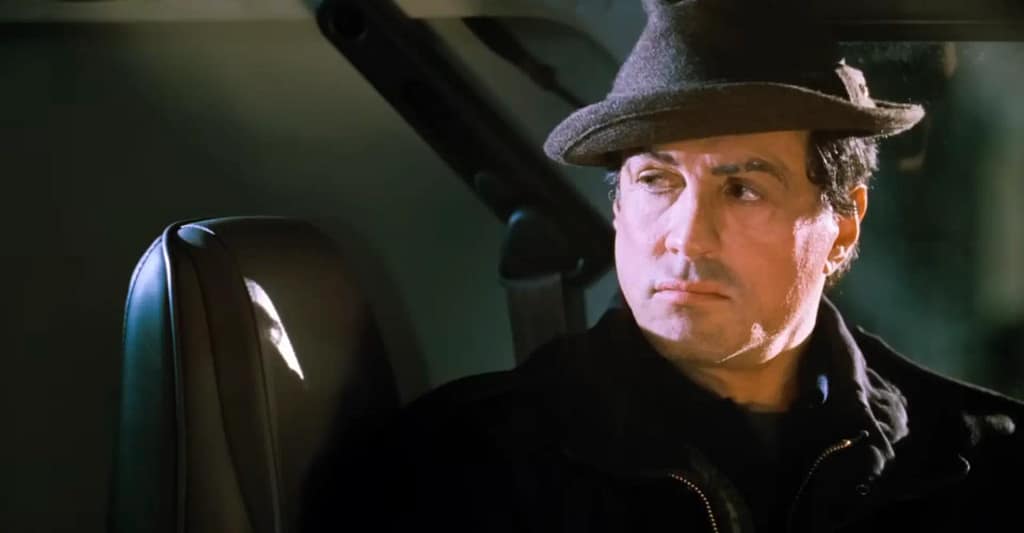 An iconic Rocky Balboa (Sylvester Stallone) wearing his iconic black fedora hat, reflecting quietly with a somber expression. Image credit: Rocky Balboa - film still via Rotten Tomatoes Official Trailer/YouTube.