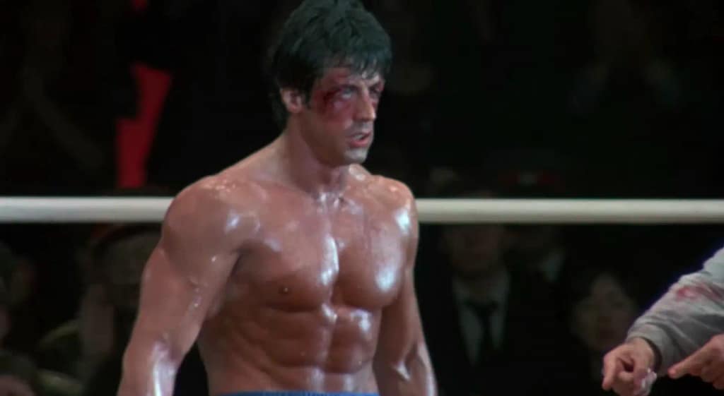 A bloody but determined Rocky Balboa (Sylvester Stallone) standing in a boxing ring showcasing physical perseverance. Image credit: Rocky IV - film still via Official Rocky Balboa/YouTube.
