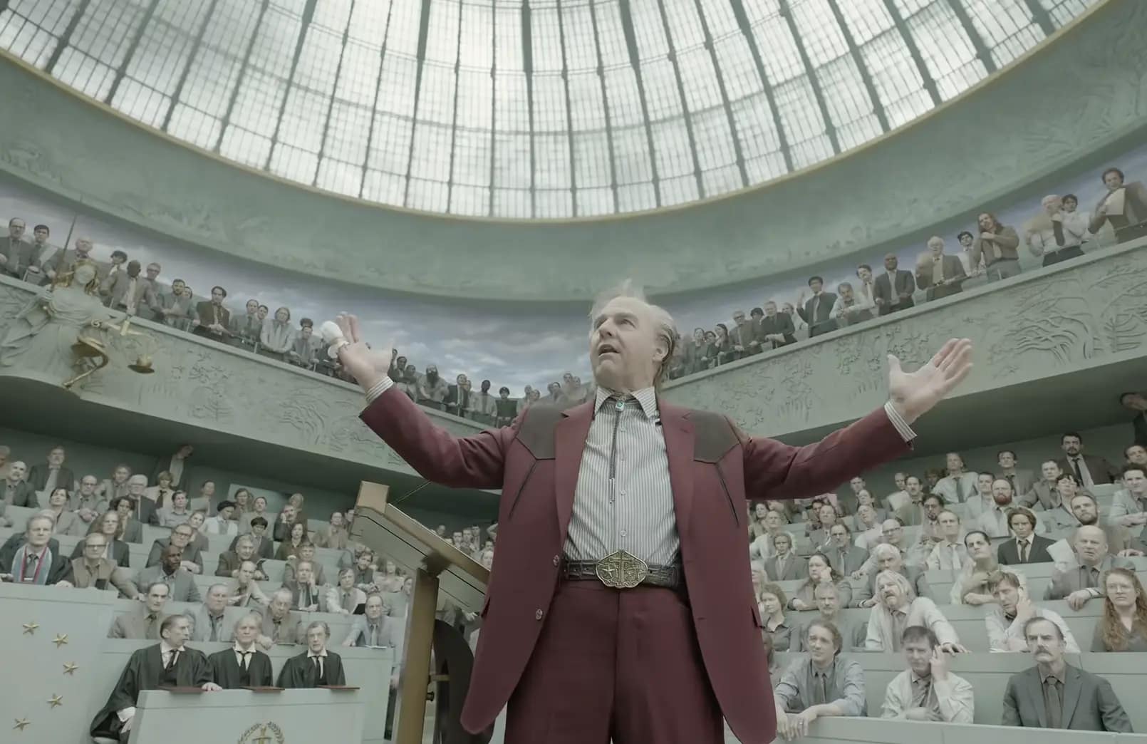 Tom Cruise as Digger Rockwell standing with arms open in a curved courtroom filled with onlookers. Image credit: Digger (2026) film still - via Warner Bros./IMAX/YouTube.